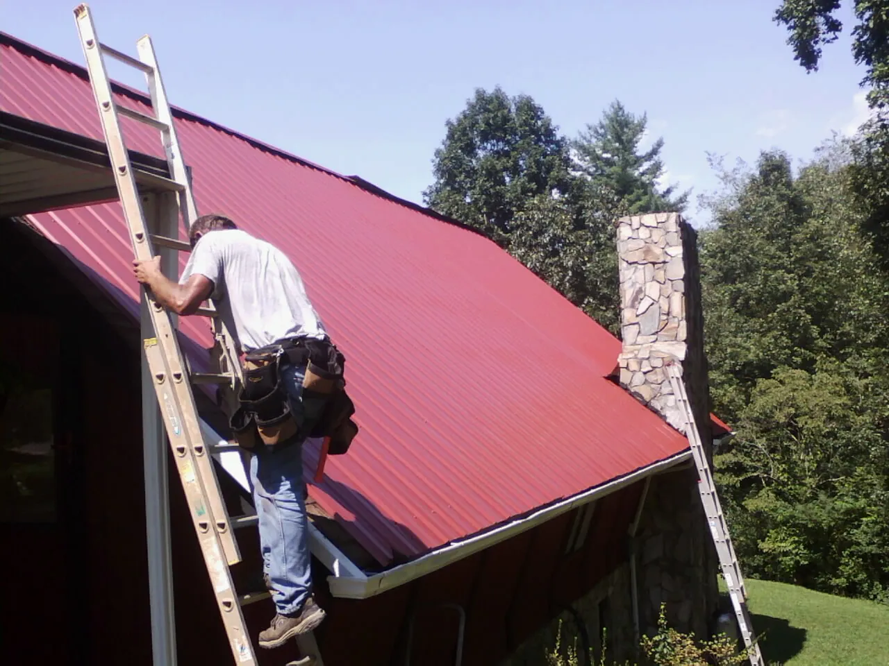 Roofer on a ladder beside a red metal roof and stone chimney — Soffit & Fascia Repair by Roofing Brothers in Bastrop